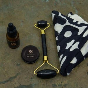 WOO luxury wellness gift set featuring lip balm, body oil, black jade roller, and stylish patterned fabric on a rustic surface, promoting sustainability and inclusive wellness.